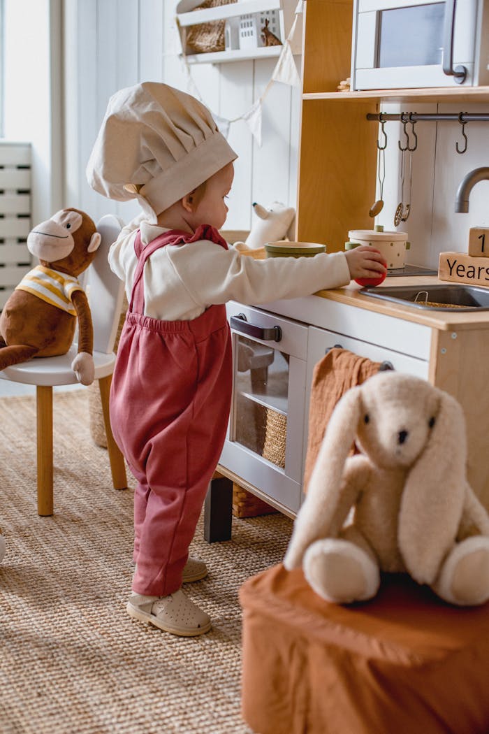 Adorable child wearing a chef hat playing in a toy kitchen with stuffed animals.